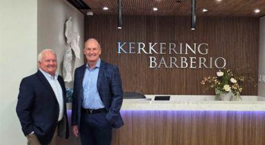 Two men in suits stand in front of a reception desk with a “Kerkering Barberio” sign on the wood-paneled wall behind them.